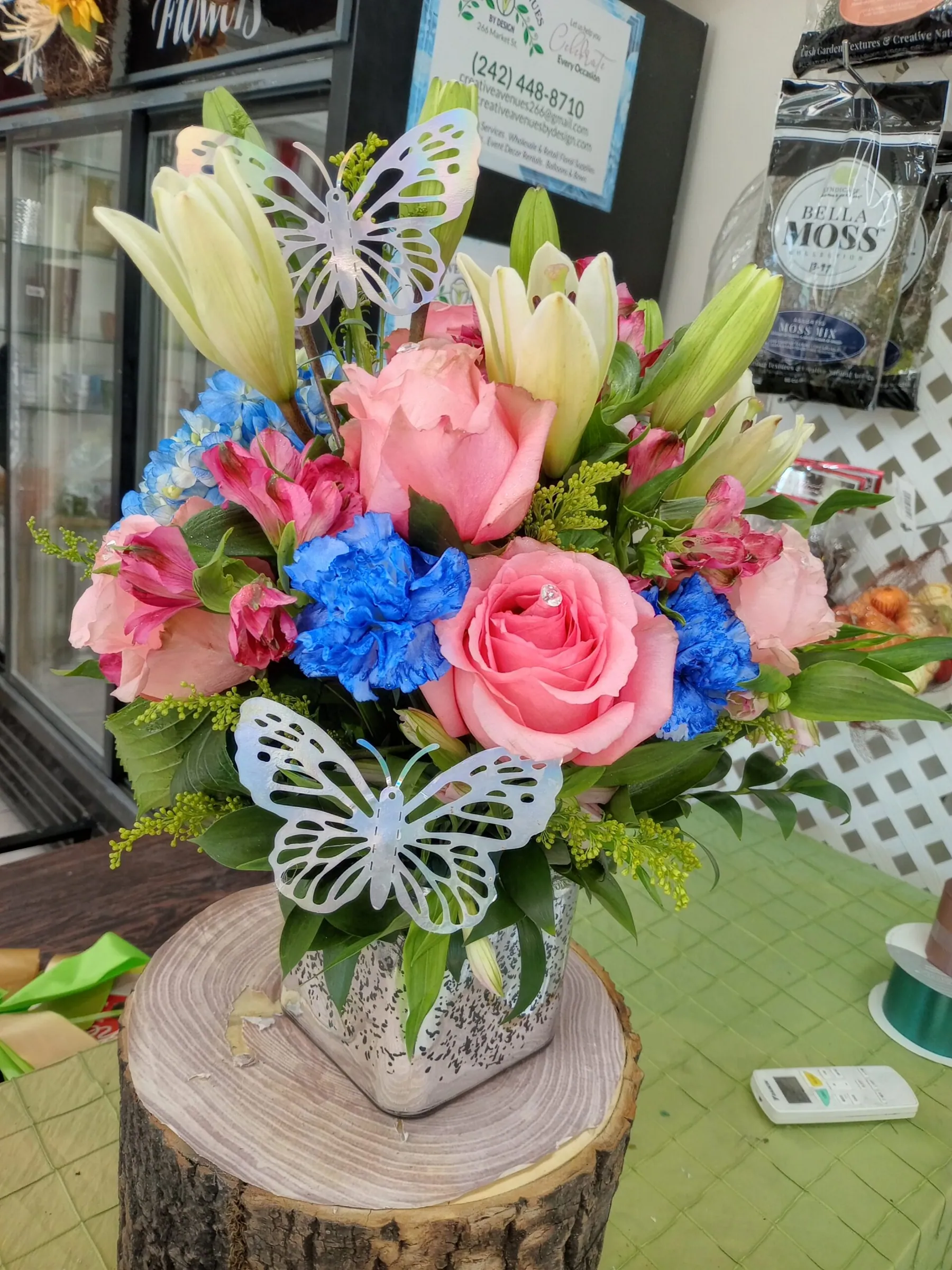 Vase arrangement of pink roses, blue blooms and lilies finished with a butterfly accent by Creative Avenues
